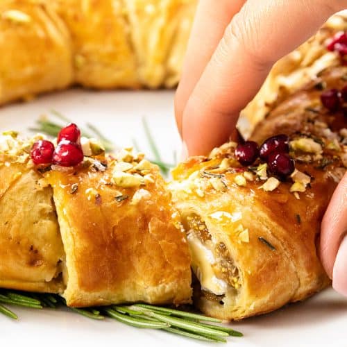 A hand with manicured nails lifts a slice of golden, flaky brie in puff pastry with jam, topped with nuts and red berries, garnished with sprigs of rosemary on a white plate.