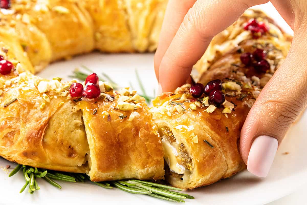 A hand with manicured nails lifts a slice of golden, flaky brie in puff pastry with jam, topped with nuts and red berries, garnished with sprigs of rosemary on a white plate.