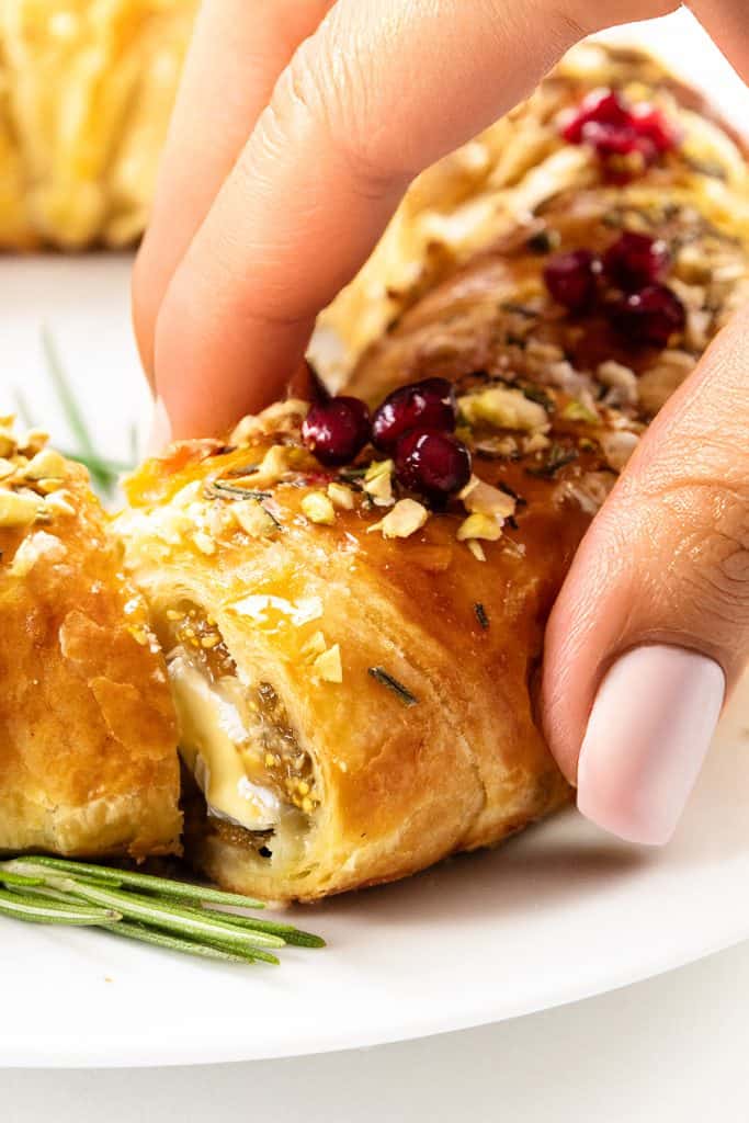A close-up of a hand picking up a slice of golden, flaky puff pastry Christmas wreath topped with nuts and red berries, revealing a creamy brie filling inside. The pastry sits on a white plate with a rosemary garnish.