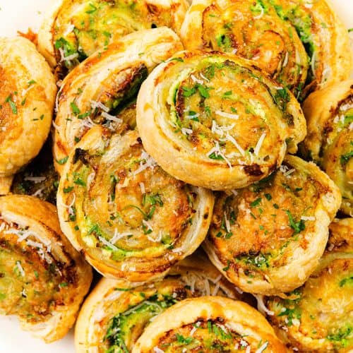 Sepiring Puff Pastry Pinwheels berwarna coklat keemasan diisi dengan bumbu hijau, keju parut, dan sentuhan Pesto Cream Cheese, ditata rapi dan ditaburi keju parut dan peterseli.