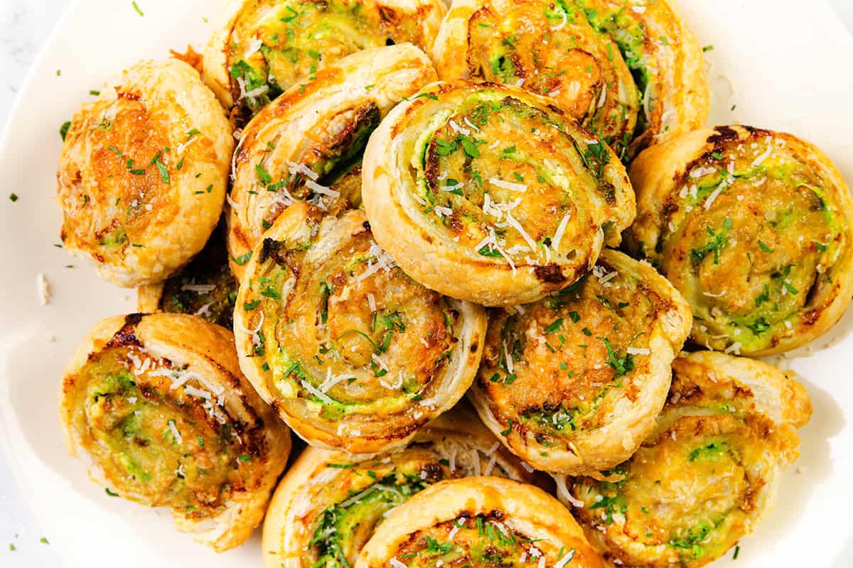 Sepiring Puff Pastry Pinwheels berwarna coklat keemasan diisi dengan bumbu hijau, keju parut, dan sentuhan Pesto Cream Cheese, ditata rapi dan ditaburi keju parut dan peterseli.