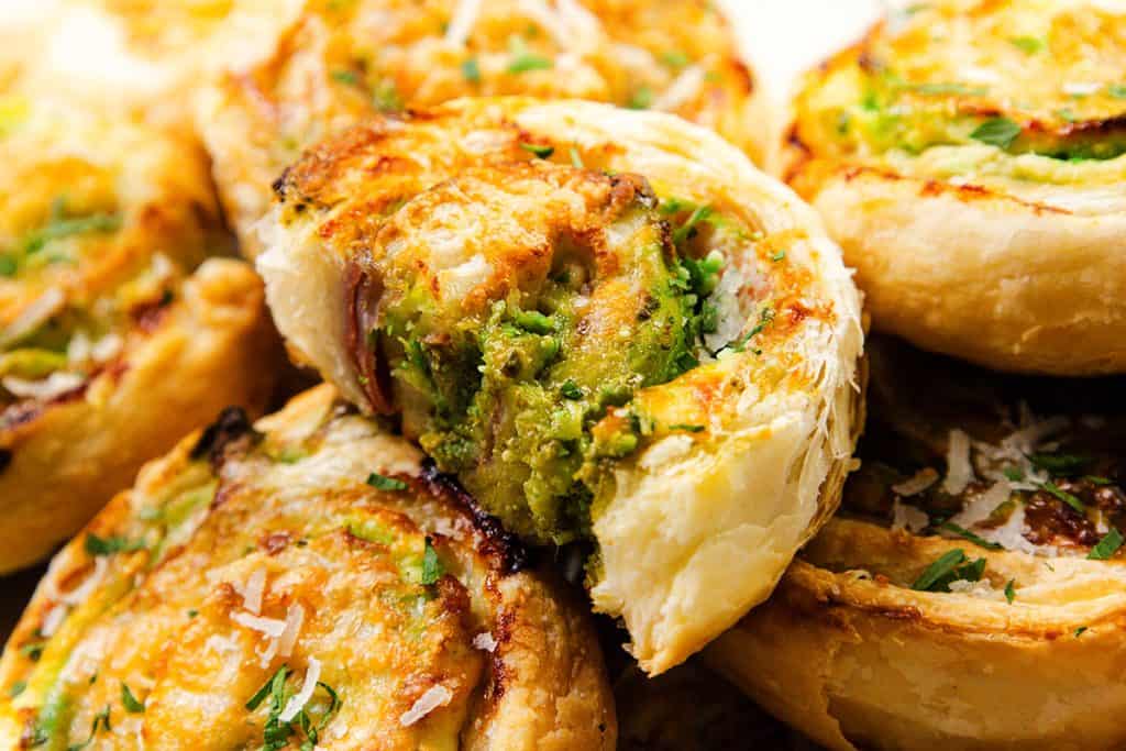 Tampilan jarak dekat dari Pinwheels Puff Pastry emas yang diisi dengan herba hijau, Keju Krim Pesto, dan di atasnya diberi keju parut dan herba segar. Kue-kue tersebut ditumpuk berdekatan, menonjolkan teksturnya yang renyah dan gurih.