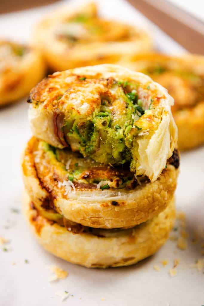 Tampilan jarak dekat dari tiga kincir puff pastry berwarna emas yang ditumpuk di atas kertas perkamen, diisi dengan herba hijau, keju krim pesto, dan mungkin sayuran atau keju, menampilkan tekstur panggang yang renyah.