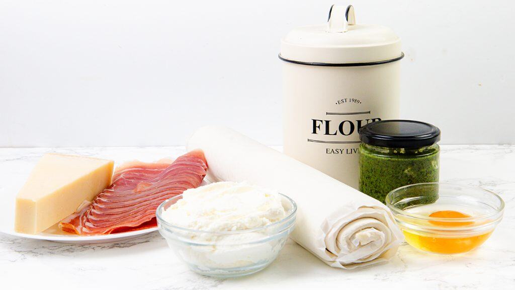 Piring dengan prosciutto dan keju Parmesan, semangkuk keju ricotta, keju krim pesto, mangkuk kaca berisi telur, selembar kue gulung yang siap untuk kincir puff pastry, dan tabung tepung di permukaan putih.