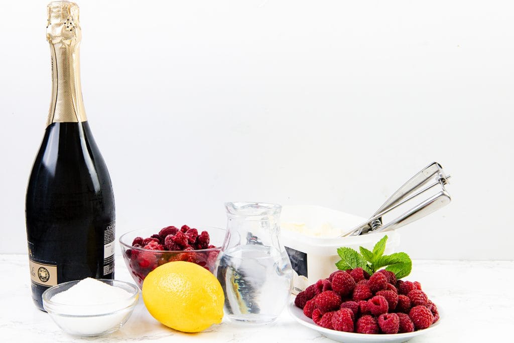 A bottle of sparkling wine, a bowl of sugar, a lemon, glass bowl of raspberries, a pitcher of water, an ice cream tub with scoop, a plate of raspberries, and mint sprigs arranged on white&mdash;perfect for making Champagne floats or other sparkling desserts.