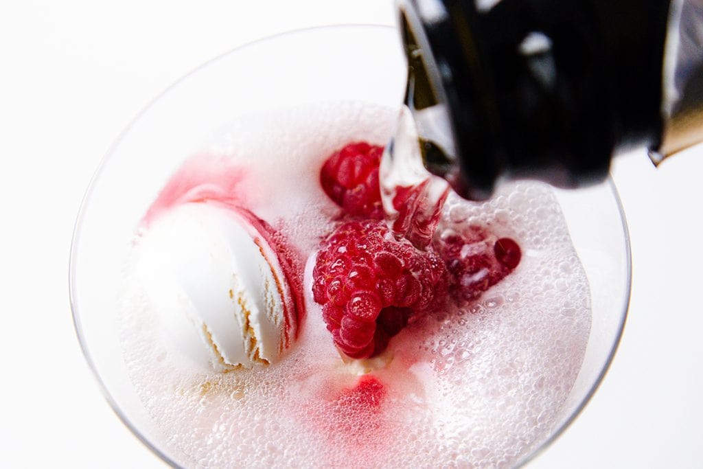 A close-up of a Champagne Float being poured into a glass containing a scoop of vanilla ice cream and fresh raspberries, creating sparkling bubbles and a foamy texture on the surface.