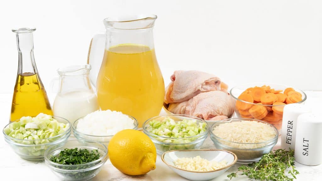 A variety of ingredients for Chicken and Rice Soup on a white surface, including a jug of broth, raw chicken pieces, sliced carrots, rice, chopped onions, celery, green onions, fresh parsley, garlic, lemon, oil, milk, fresh herbs, and salt and pepper shakers.