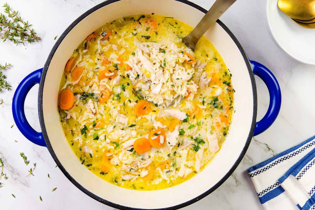 A pot of Chicken and Rice Soup with sliced carrots, herbs, and a creamy broth. A ladle rests in the pot, while fresh thyme and a kitchen towel are nearby on a white marble surface.