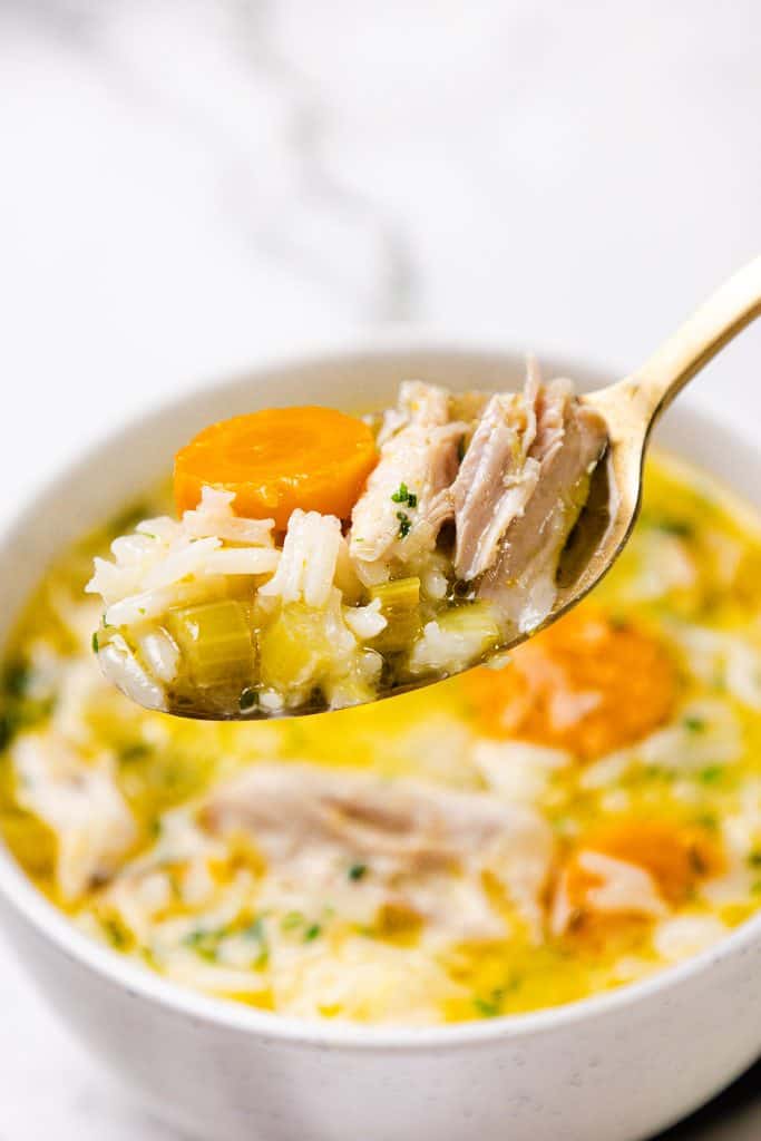 A close-up of a spoonful of Chicken and Rice Soup with tender chicken, rice, carrots, and celery held above a bowl brimming with the comforting soup.