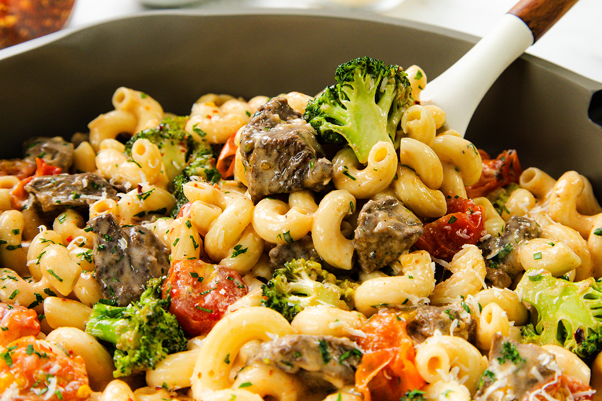A close-up of creamy steak pasta with tender beef chunks, broccoli florets, and tomato pieces, garnished with herbs, being stirred in a pan with a white-handled spoon.