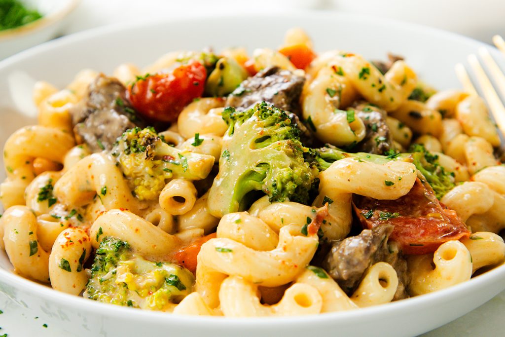 A close-up of a plate of Creamy Steak Pasta with broccoli, cherry tomatoes, and pieces of meat, garnished with chopped parsley.