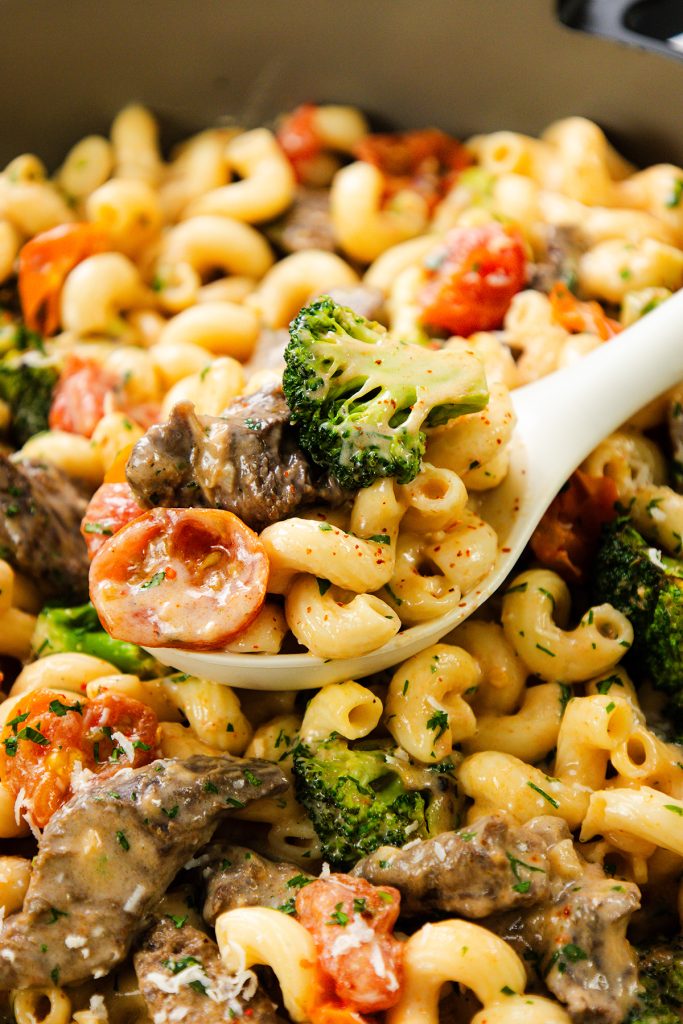 Close-up of Creamy Steak Pasta featuring elbow macaroni, sliced beef, broccoli, and cherry tomatoes, garnished with herbs and served with a white spoon.