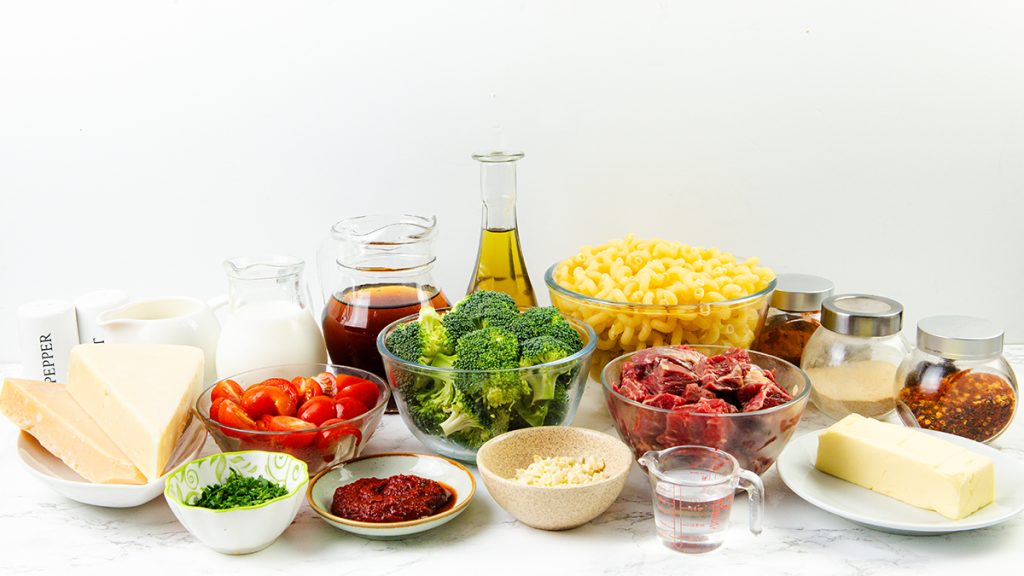 A selection of ingredients on a white surface, including pasta, broccoli, raw beef for steak pasta, cherry tomatoes, parmesan cheese, butter, oil, milk, spices, tomato paste, broth, and chopped herbs in various bowls and containers.