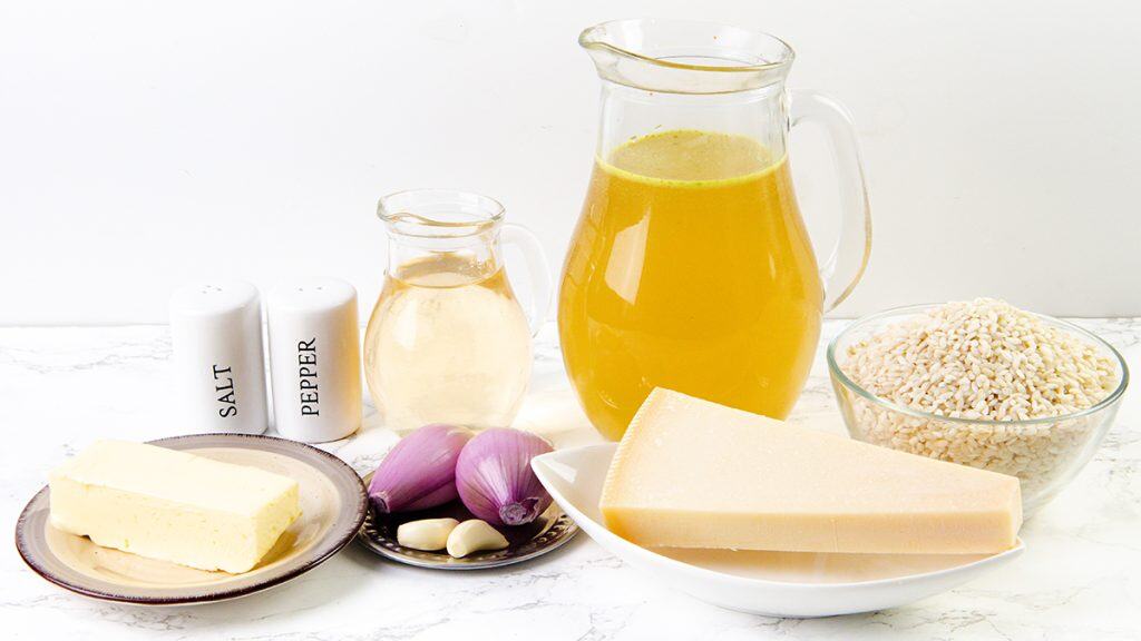 A pitcher of broth, a jug of white wine, a bowl of uncooked rice, Parmesan for grating—everything you need for Parmesan Risotto—plus butter, shallots, garlic cloves, and salt and pepper shakers on a white surface.