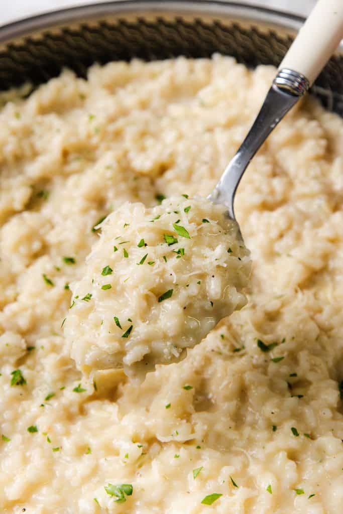 A close-up of a spoonful of creamy Parmesan Risotto garnished with finely chopped herbs, held above a pan filled with more easy risotto.