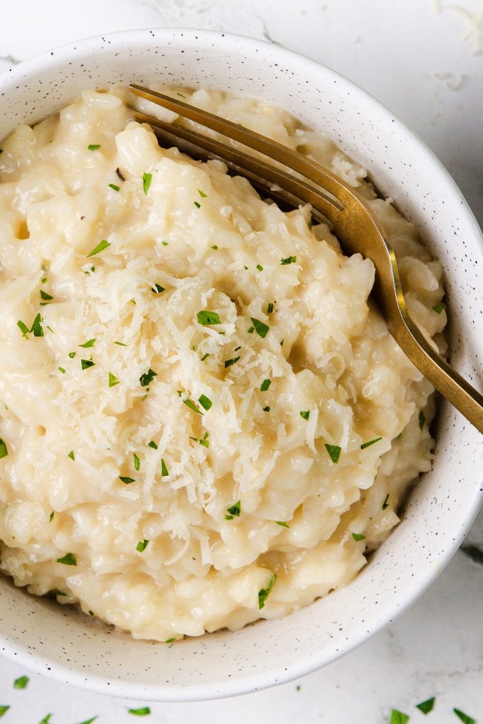 A close-up of creamy Parmesan Risotto in a white bowl, topped with grated cheese and chopped parsley, with a gold fork resting on the side. This easy risotto is perfect for a cozy dinner.