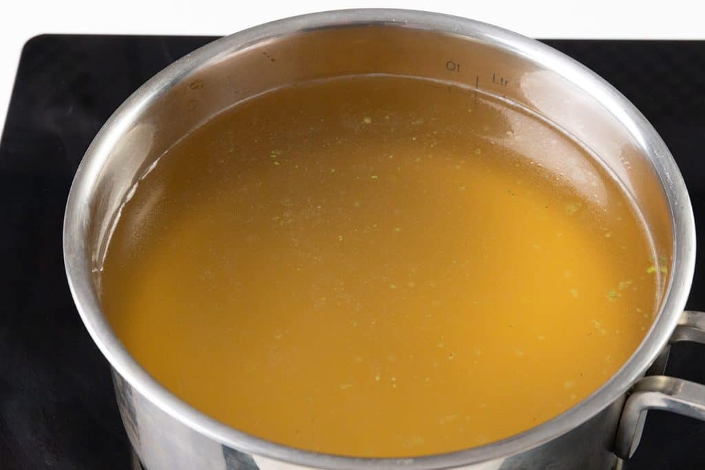 A stainless steel pot filled with golden-brown broth sits on a black stovetop, ready to become an easy risotto. The surface of the broth is clear with a few tiny green flecks visible.