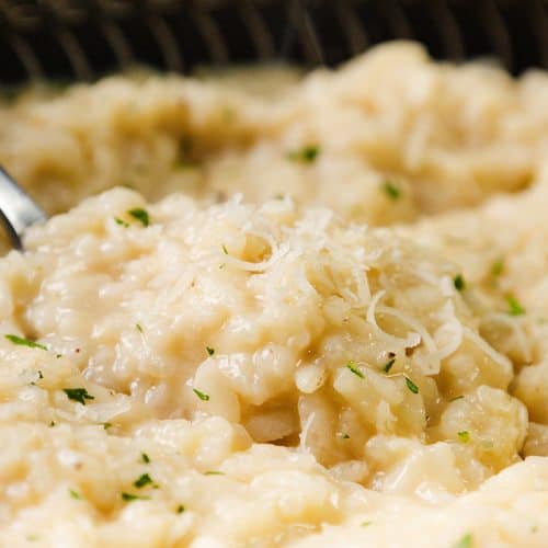 A close-up of creamy Parmesan Risotto garnished with grated cheese and chopped herbs, with a spoon partially submerged in the dish.