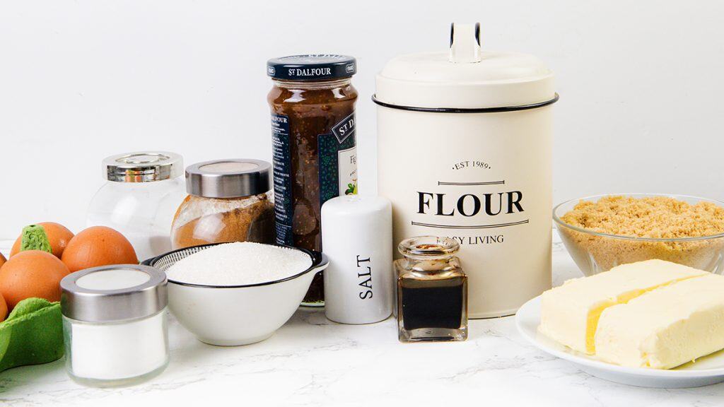 A collection of baking ingredients on a white counter, perfect for making Fig Filled Cookie Bars, including eggs, sugar, brown sugar, flour in a canister, butter, salt, spices, vanilla extract, baking powder, and a jar of jam or preserves.