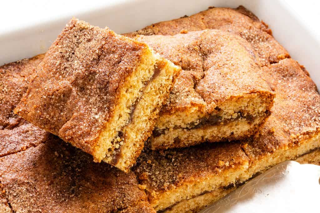 Golden, cinnamon-sugar snickerdoodle cookie bars are stacked in a baking dish. The bars have a soft, cake-like texture with a visible cinnamon swirl and a slightly crisp, sugary topping.