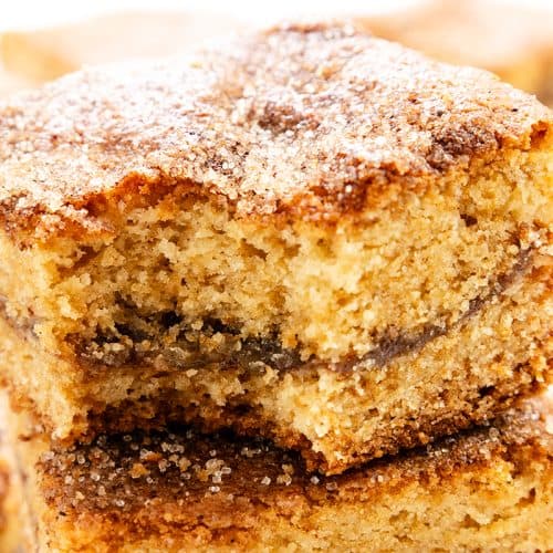Two cinnamon sugar bars are stacked, showing their soft, crumbly texture and a layer reminiscent of classic Fig Filled Cookie Bars. More bars are blurred in the background. The bars appear moist and golden brown.
