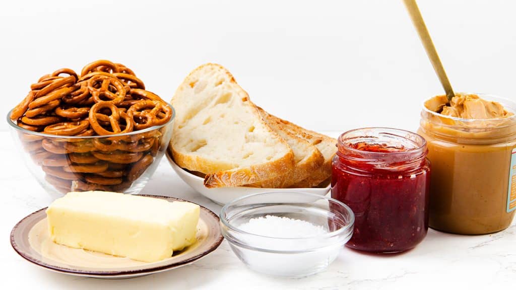 A variety of snack ingredients on a white surface: a bowl of pretzels with a crunchy pretzel coating, slices of bread, a jar of peanut butter and jelly with a spoon, a dish of butter, and a small bowl of salt.