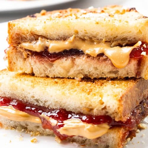 Two halves of a Grilled Peanut Butter and Jelly sandwich are stacked on a white surface, showing layers of melted peanut butter and red jelly between toasted, golden-brown bread slices.