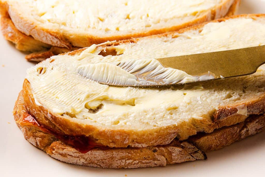 A slice of bread is being spread with butter using a knife, with another slice and a bit of red jam visible&mdash;perfect for making a classic Peanut Butter and Jelly sandwich.