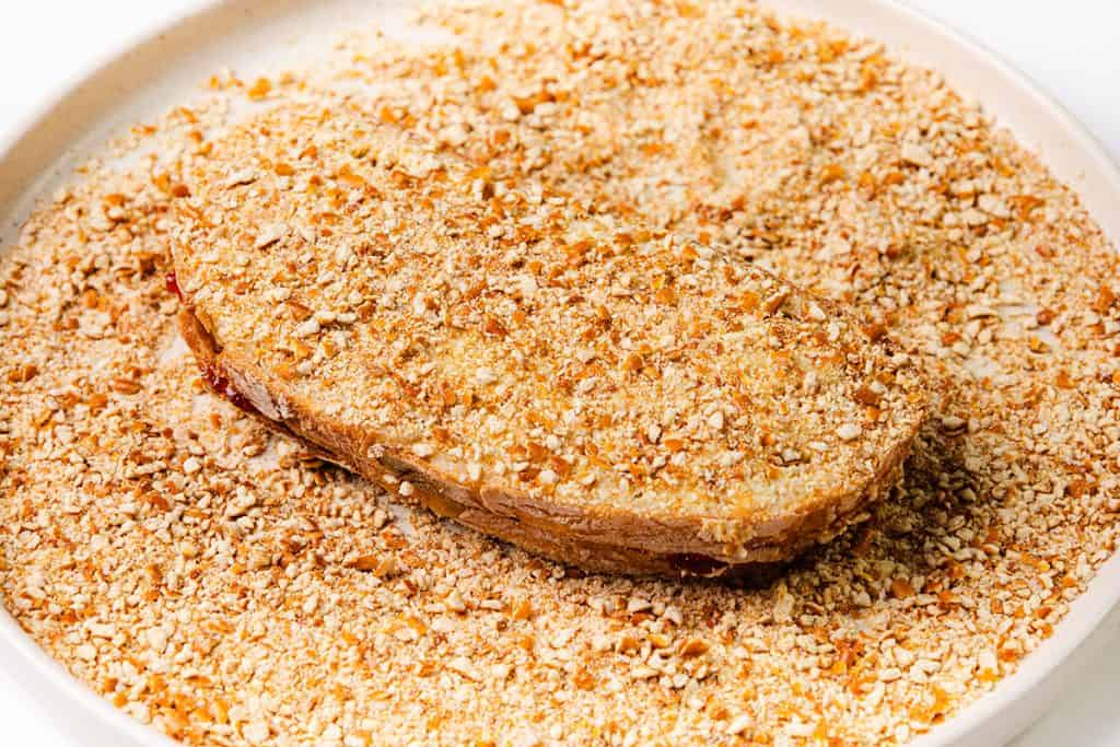A sandwich coated with a crunchy pretzel coating sits on a plate filled with more crumbs, ready for further preparation or cooking.