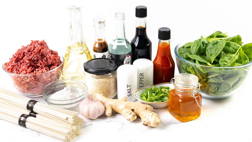 A variety of ingredients for a Beef Noodle Recipe are arranged on a white surface, including ground beef, noodles, spinach, garlic, ginger, green onions, sesame seeds, sauces, salt, pepper, oil, honey, and cornstarch in bowls and jars.