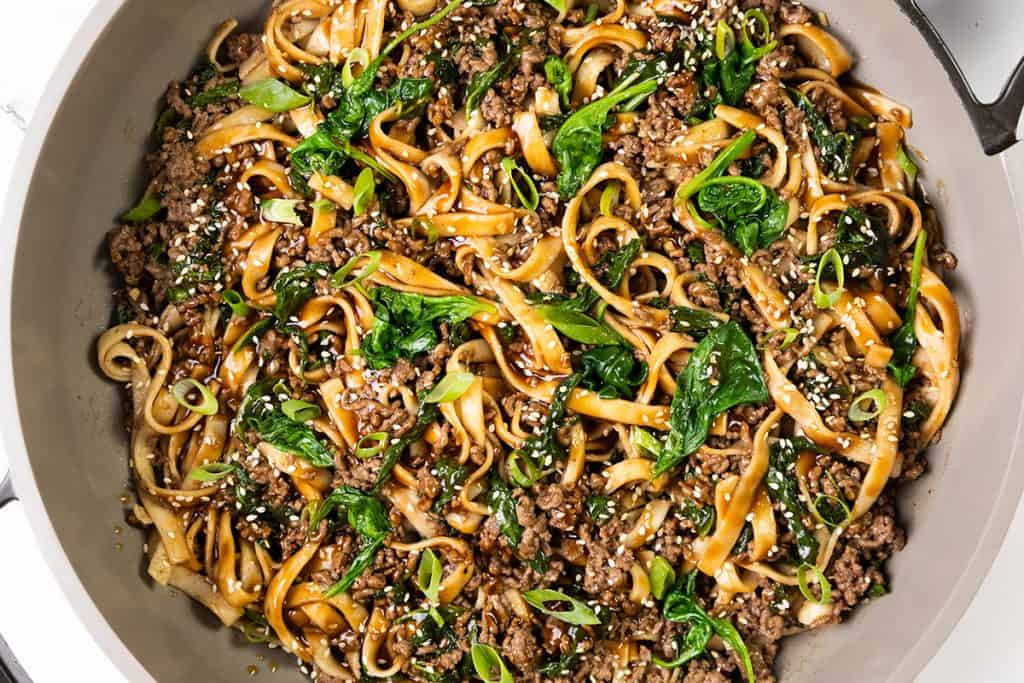 A skillet filled with stir-fried sesame noodles, ground beef, spinach, green onions, and sesame seeds in a savory sauce.