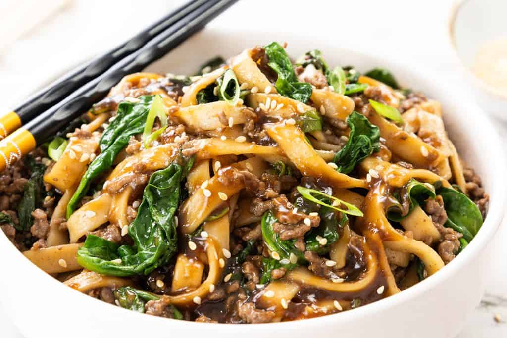 A bowl of sesame noodles with ground beef, spinach, and scallions, topped with sesame seeds. Chopsticks rest on the bowl’s edge. The dish is coated in a glossy brown sauce for a delicious beef noodle recipe.