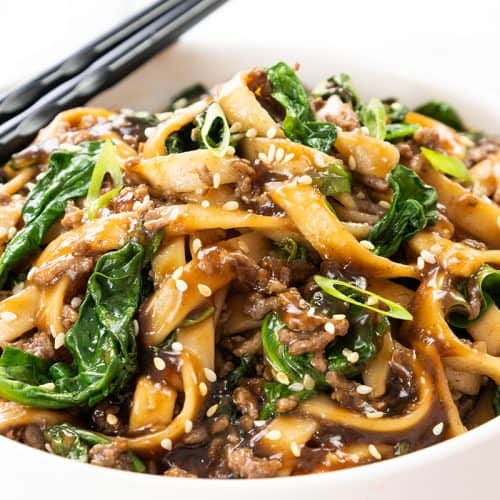 A bowl of sesame noodles with ground beef, spinach, and scallions, topped with sesame seeds. Chopsticks rest on the bowl&rsquo;s edge. The dish is coated in a glossy brown sauce for a delicious beef noodle recipe.