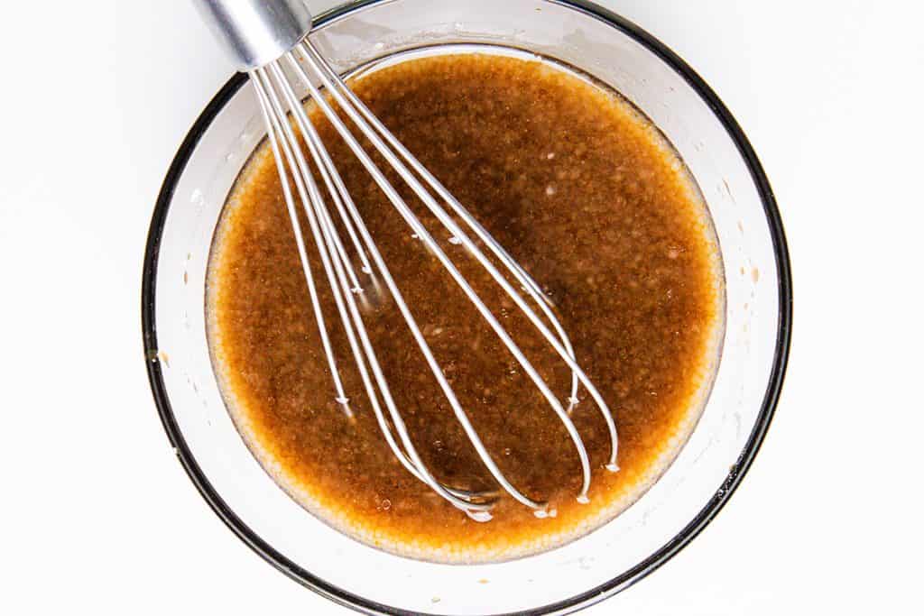 A metal whisk rests in a glass bowl filled with a brown, grainy liquid mixture for sesame noodles, viewed from above on a white background.