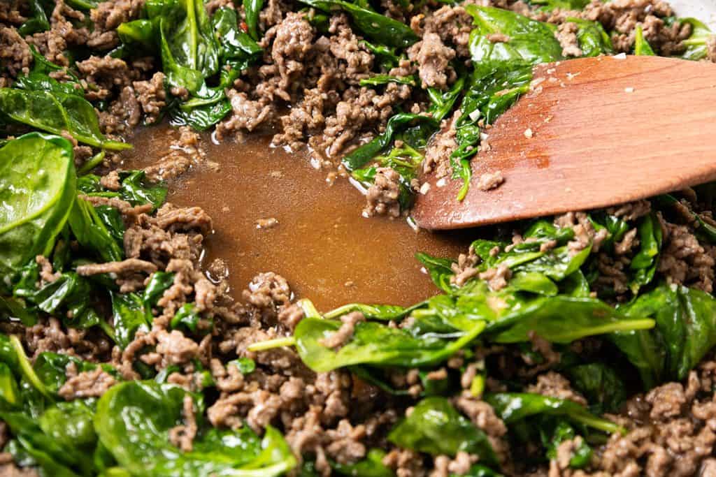 Close-up of ground beef and wilted spinach in a pan, with a pool of sauce in the center and a wooden spoon stirring&mdash;perfect for adding to your favorite Beef Noodle Recipe or tossing with sesame noodles.