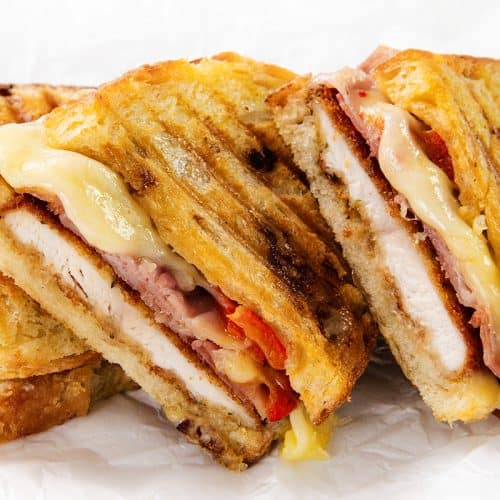 Three grilled Italian Chicken Cutlet panini sandwiches filled with melted cheese, roasted red peppers, bacon, and tender chicken breast slices, stacked closely together on white parchment paper.