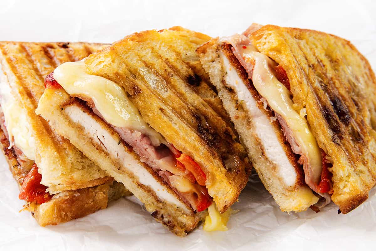 Three grilled Italian Chicken Cutlet panini sandwiches filled with melted cheese, roasted red peppers, bacon, and tender chicken breast slices, stacked closely together on white parchment paper.