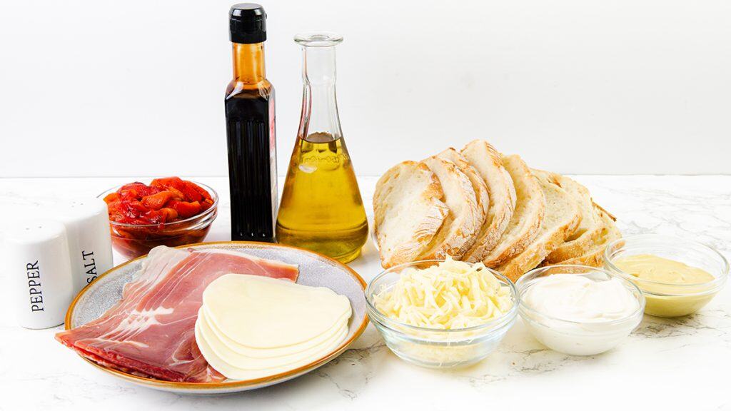 Assorted ingredients on a white surface, perfect for creating an Italian Sandwich—sliced bread, prosciutto, provolone cheese, shredded cheese, roasted red peppers, oil, balsamic vinegar, mayonnaise, mustard, and salt and pepper shakers.