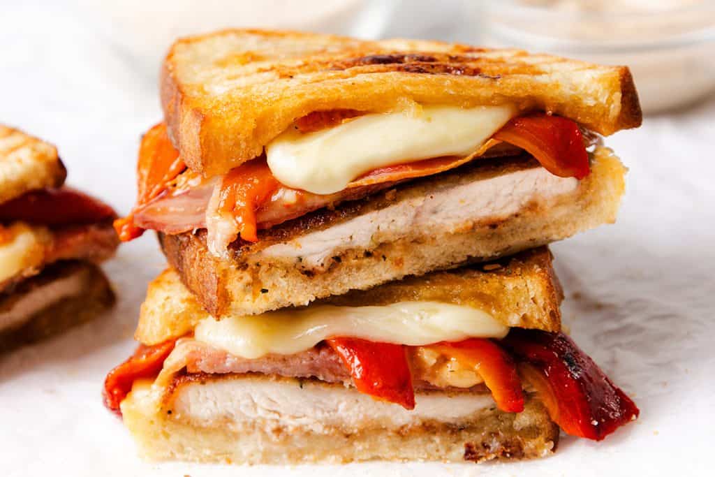 Two stacked grilled Italian sandwiches with golden toasted bread, filled with melted cheese, red bell peppers, ham, and a thick slice of Italian chicken cutlet. The melted cheese and peppers are visible at the edges.