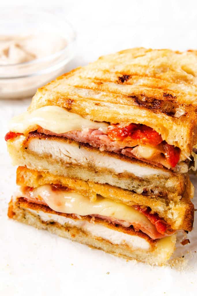 A close-up of an Italian Chicken Cutlet sandwich cut in half, showing layers of melted cheese, roasted red peppers, breaded chicken cutlet, and crispy bacon between toasted bread. A bowl of dipping sauce is blurred in the background.