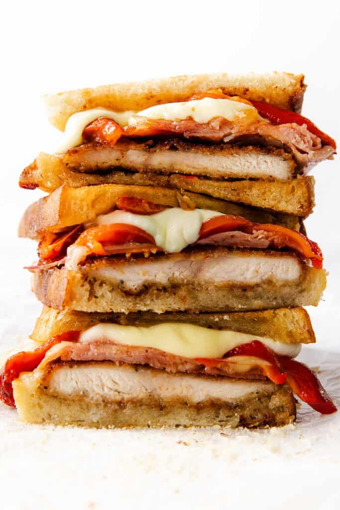 A close-up of a stacked Italian Chicken Cutlet sandwich with layers of toasted bread, crispy bacon, melted cheese, roasted red peppers, and thick slices of grilled chicken, all on a white background.