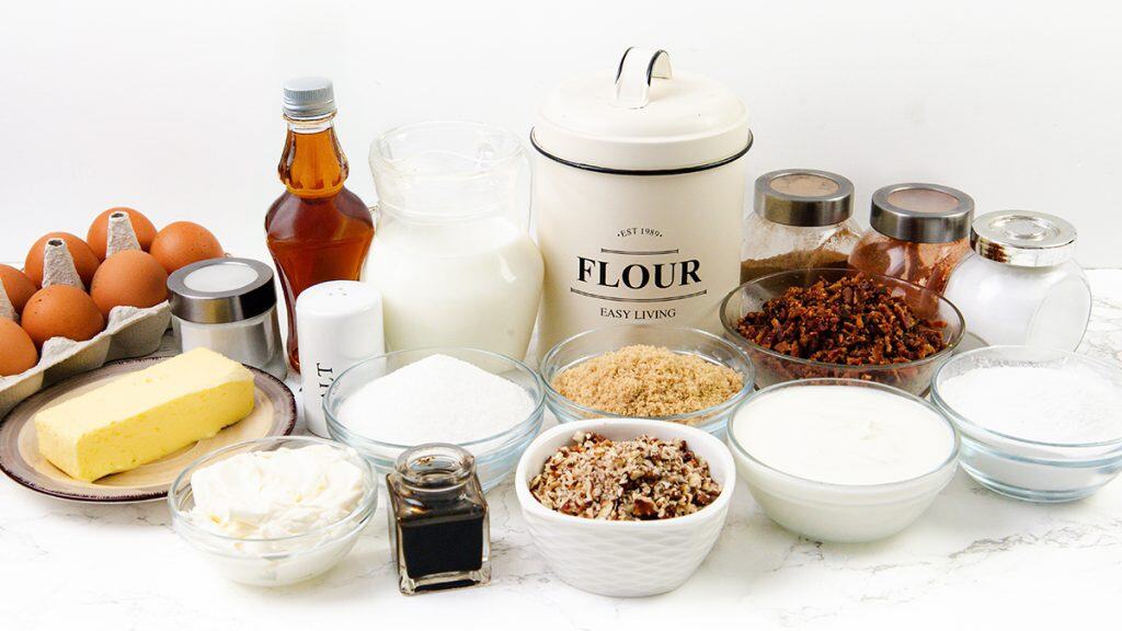 A variety of baking ingredients are displayed on a white surface, ready to be used in a Maple Bacon Bundt Cake, including eggs, butter, flour, milk, sugar, vanilla extract, pecans, baking powder, baking soda, salt, sour cream, and brown sugar.