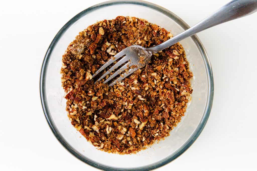 A glass bowl filled with a crumbly mixture of chopped nuts and spices for a Maple Bacon Cake, with a metal fork resting inside, on a white background.
