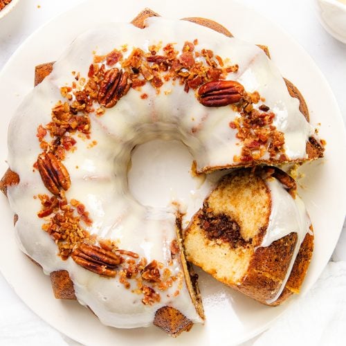 A Maple Bacon Bundt Cake with white glaze, topped with pecans and crumbled maple bacon, sits on a white plate. A slice is cut and slightly separated, revealing a cinnamon swirl inside the cake.
