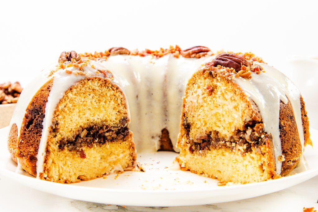 A bundt cake with a creamy white glaze, topped with pecans and crumbs. The cake has a visible inner layer of pecan filling and a slice missing, revealing its moist, golden interior&mdash;perfect for maple bacon cake lovers.