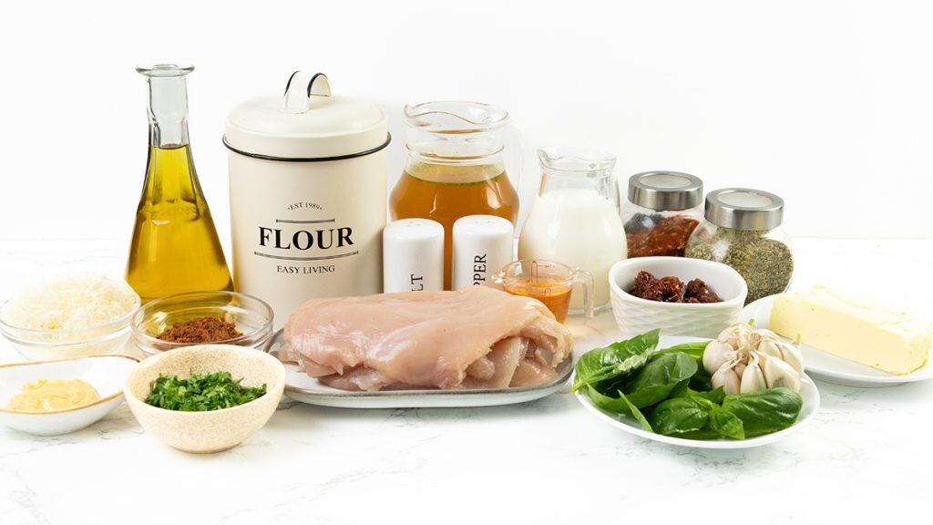 A variety of ingredients on a white surface, including raw chicken breasts for a creamy chicken recipe, spinach leaves, garlic, butter, spices in jars, shredded cheese, fresh herbs, oil, flour canister, broth, heavy cream, and condiments.