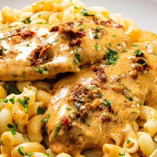 Creamy sun-dried tomato chicken recipe served over curly pasta, garnished with chopped herbs on a white plate. A gold fork and spoon rest beside this inviting Marry Me Chicken dish.