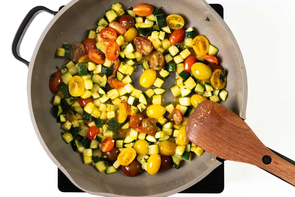 A frying pan on a stovetop with chopped zucchini and halved cherry tomatoes being sautéed, stirred with a wooden spatula—perfect for pairing with Mediterranean chicken.
