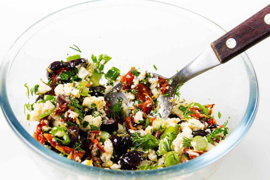 A glass bowl filled with a Mediterranean salad made of chopped herbs, feta cheese, Kalamata olives, sun-dried tomatoes, and green onions sits beside savory Mediterranean Chicken. A metal spoon with a wooden handle rests in the bowl.