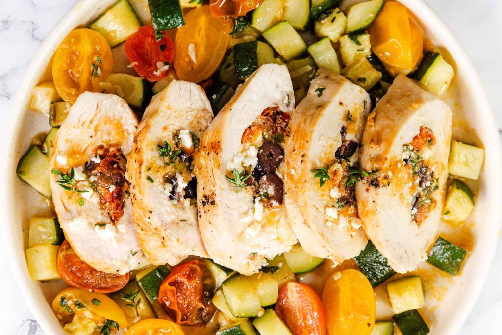 Sliced Mediterranean Stuffed Chicken breast filled with cheese, olives, and sun-dried tomatoes, served on a plate with diced zucchini and colorful cherry tomatoes.